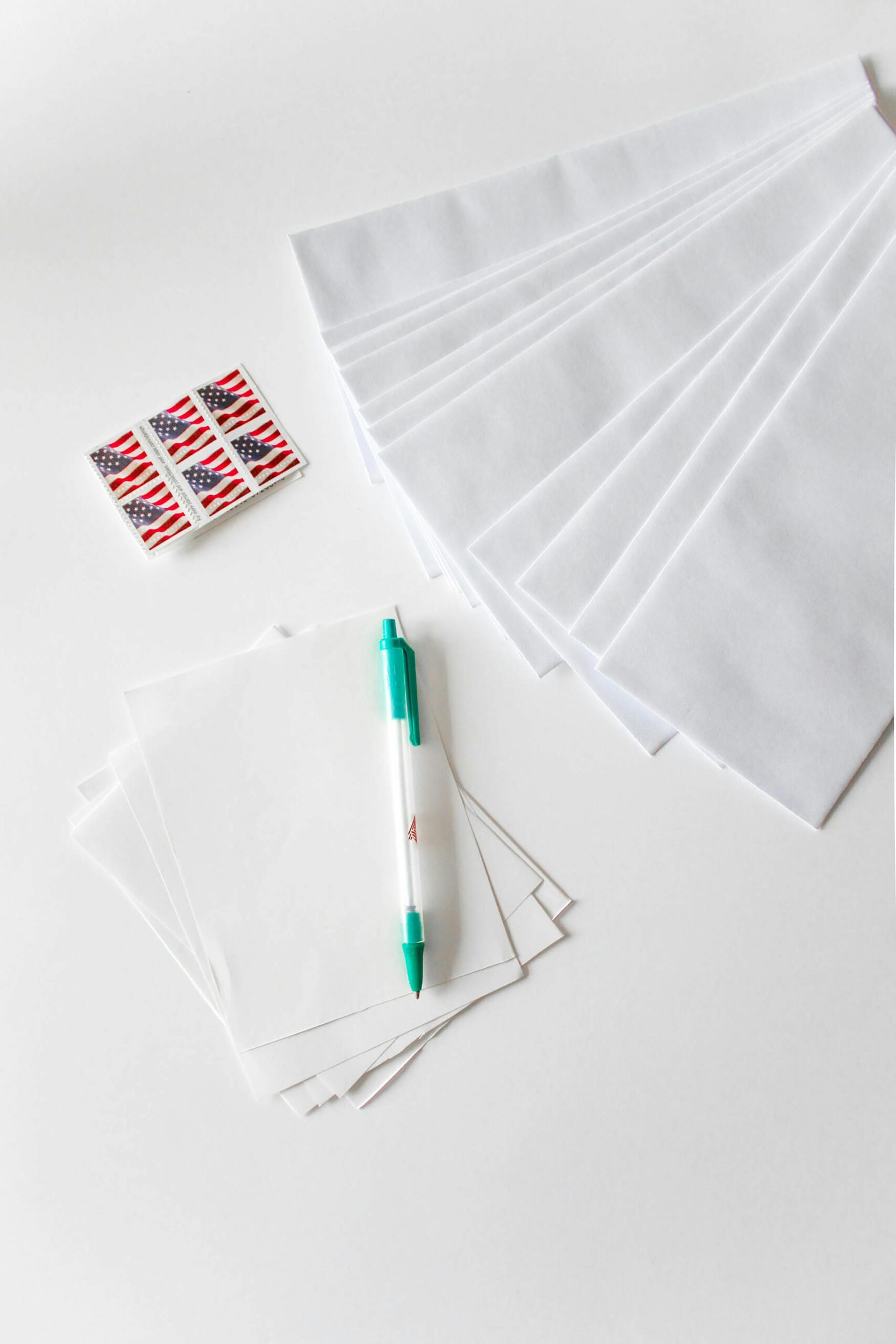 Flat lay of envelopes, blank paper, pen, and American flag stamps on white background.