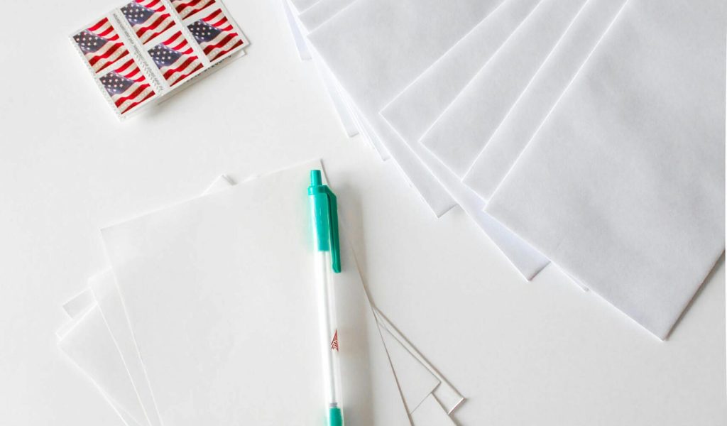 Flat lay of envelopes, blank paper, pen, and American flag stamps on white background.