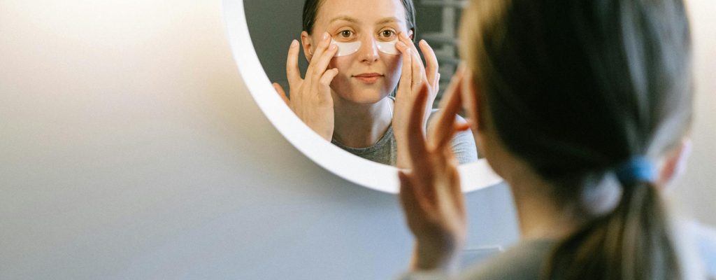 A woman applying under-eye patches while looking in a round wall mirror, focusing on skincare.