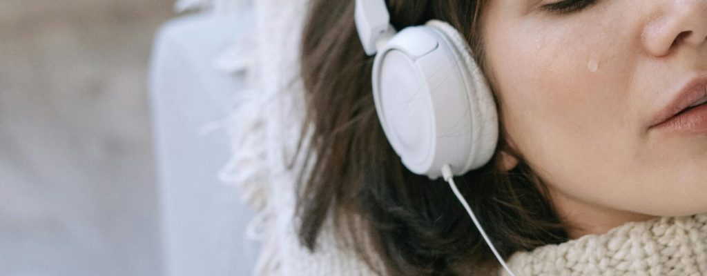Young woman lying down indoors, wearing headphones and a knitted sweater, with tears on her face.